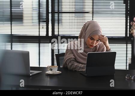 Nachdenkliche oder stressige islamische Mädchen in Hijab fühlen sich von der Arbeit vor dem Tablet-Computer auf dem Schreibtisch im Büro aufgebracht. Stockfoto