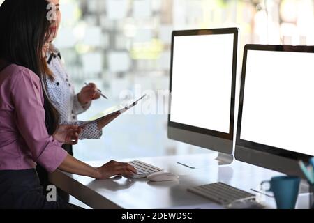 Geschäftsleute arbeiten zusammen und sprechen durch neue Ideen, während sie vor mehreren Computerbildschirmen stehen. Stockfoto