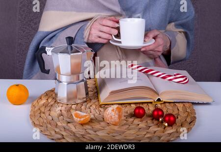 Nahaufnahme Stillleben mit einem Buch, eine Geysir-Kaffeemaschine, Weihnachtsdekor auf dem Tisch. Eine Frau hält eine weiße Tasse mit einem heißen Kaffeegetränk. Selektiver FOC Stockfoto