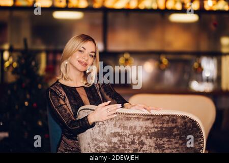 Eine blonde Frau in einem schwarzen Kleid lächelt und schaut auf die Kamera auf dem Bokeh-Hintergrund des neuen Jahres. Weihnachten, Urlaub