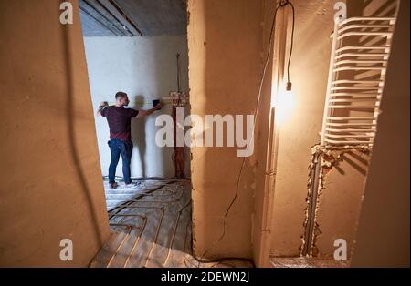 Rückansicht auf jungen Techniker in leerer unvollendeter Wohnung, die die Wand mit Hilfe von Maßband misst, Blick durch den Eingang vom Flur. Konzept der Renovierung und Restaurierung. Stockfoto