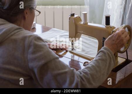 Eine Frau näht Stoffvorhänge mit Hilfe einer antiken Handnähmaschine in einem Raum eines Hauses in der Nähe des Fensters. Nähausrüstung mit zwei Spulen Stockfoto