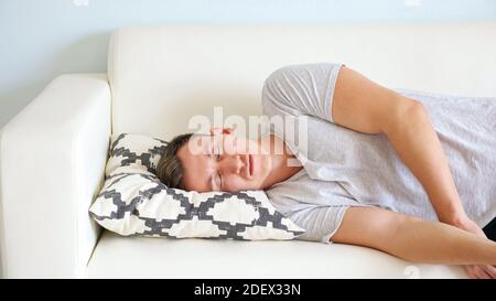 Müde junger Mann mit geschlossenen Augen in weißem T-Shirt liegt Auf weicher Couch mit kleinem Designer-Kissen im hellen Raum Nahaufnahme zu Hause Stockfoto