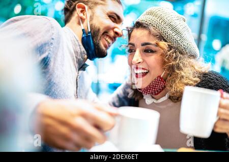 Glückliches Paar mit Gesichtsmaske Spaß zusammen an der Bar Cafeteria - Neue normale Lifestyle-Konzept mit jungen Menschen reden Und amerikanischen Kaffee trinken