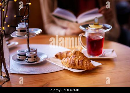 Ein kurzer Schuss Kaffeetisch mit einer Tasse Tee und Croissant, während eine Frau ein Buch in der Hand hält und liest. Stockfoto