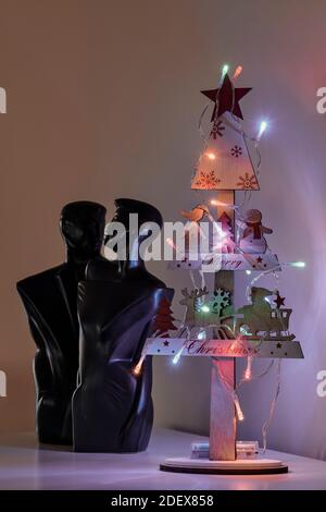 Miniatur-Weihnachtsbaum aus Holz mit Weihnachtsmann und einem Schneemann geschmückt, beleuchtet mit farbigen Lichtern und der Skulptur eines verliebten Paares Stockfoto