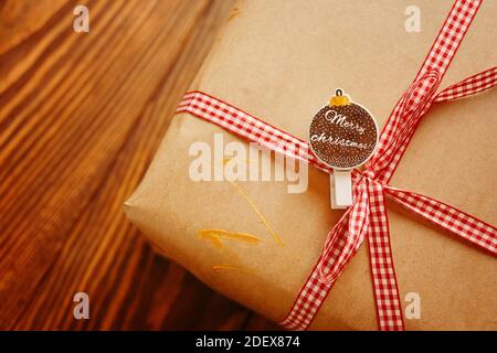 Geschenkbox auf Holztisch. Dekorative Wäscheklammer in Form von Kugel mit fröhlichen Weihnachten Kalligraphie Inschrift. Bastelpapier mit kariertem rotem Band. Konzept von Neujahr und Weihnachten. Stockfoto