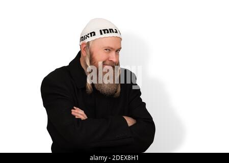 Listig bärtiger jüdischer Mann lächelt listig und blickt auf die Kamera über weißem Hintergrund. Charmanter jüdischer Mensch mit Seitenschwänzen in weißem Kippah Stockfoto