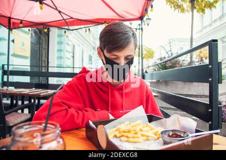 Das Tragen von Masken an öffentlichen Orten. Der junge Mann sitzt in einer Schutzmaske in einem Straßencafé und sieht Pommes frites an. Stockfoto
