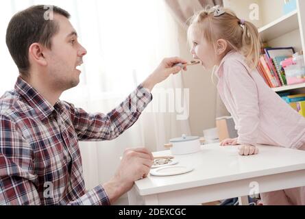 Fürsorglicher Vater füttert seine schöne Tochter mit leckerem Essen mit einem Löffel in einem gemütlichen Haus. Stockfoto