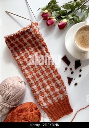 Fragment aus beigefarbenem Pullover, gestrickter Ärmel aus warmer Schafwolle. Weißer Cappuccino-Becher, getrocknete Rosen, Garnknäuel Stockfoto