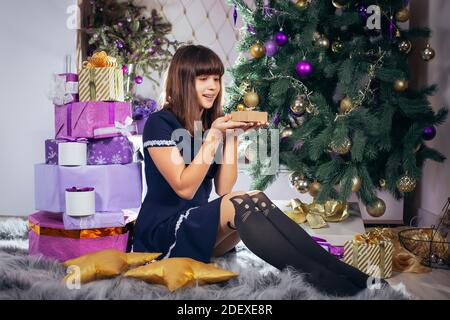 Fröhliche Teenager-Mädchen mit einem Geschenk in den Händen sitzt in der Nähe eines Weihnachtsbaums. In der Nähe ein Stapel von lila farbigen Geschenken und festliche Neujahrsdekorationen Stockfoto