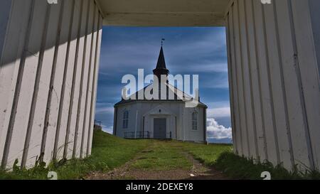 Schöne alte Holzkirche im achteckigen Stil mit weißer Fassade umgeben von grünem Gras durch ein Holztor in der Nähe von Dverberg, Norwegen. Stockfoto