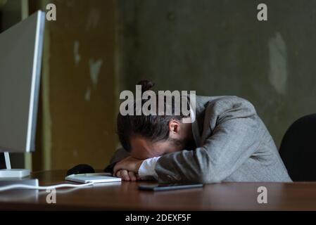 Junger bärtiger Geschäftsmann in einem Geschäftsanzug, der am Computertisch im Büro schläft. Junger Business Kerl liegend Gesicht nach unten auf dem Tisch gelangweilt jung Stockfoto