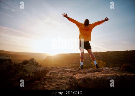 Rückansicht des jungen Mannes, der mit ausgestreckten Armen am Berggipfel während des Morgensonnenaufgangs steht. Widrigkeiten durch Motivation und überwinden Stockfoto