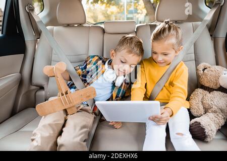 Lächelndes Mädchen hält digitale Tablette in der Nähe Bruder mit Spielzeug Flugzeug Im Auto Stockfoto