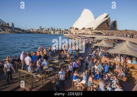 Ein geschäftiges Hafengebiet von Sydney, das zum Opernhaus führt, während Einheimische und Touristen in der Opera Bar Getränke genießen und Kontakte knüpfen. Stockfoto