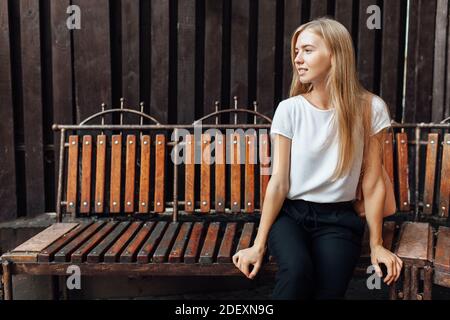 Junge schöne Mädchen, in einem weißen T-Shirt, sitzt auf einer Bank gegen eine hölzerne Laubwand Stockfoto