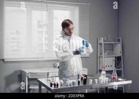 Ein Wissenschaftler in Schutzbrille und weißen Overalls, der am Tisch im Wissenschaftslabor steht und an etwas arbeitet. Stockfoto