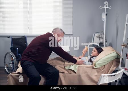 Ein älterer Mann jubelte seine Enkelin, die auf einer Krankenhausstation im Bett liegt, und brachte ihr ein Eis. Stockfoto