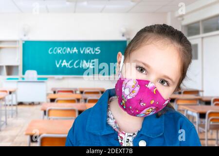 Mädchen in einem Klassenzimmer trägt eine Gesichtsmaske und die Worte Corona-Virus-Impfstoff auf einer Tafel geschrieben. Stockfoto