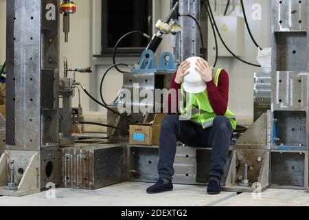 Porträt eines männlichen Fabrikmanagers in einem weißen Harthut und rotem Pullover, der seinen Kopf hält. Lösung von Werksproblemen. Arbeiter verlor den Job. Arbeitslosigkeit . Hochwertige Fotos Stockfoto