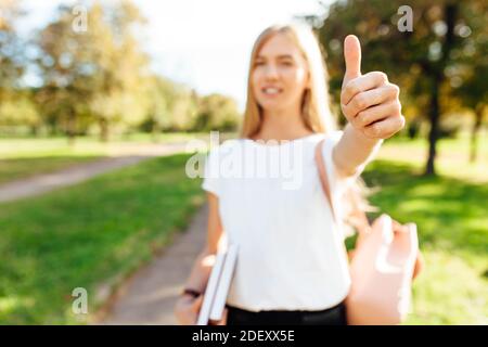 Porträt eines schönen Studenten Mädchen durch den Park zu Fuß Halten Sie Bücher in den Händen und zeigen Sie ihren Daumen nach oben Stockfoto