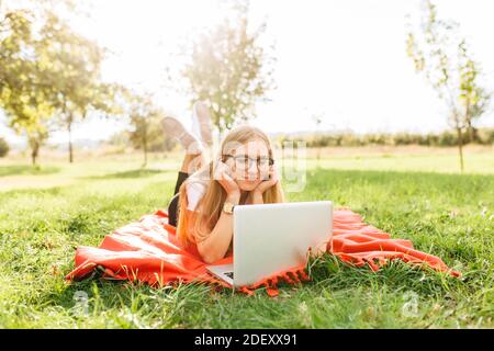 Bild einer schönen Studentin mit Brille, die auf einem Laptop in guter Laune im Park arbeitet, auf einer Decke liegend Stockfoto