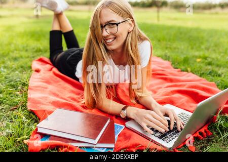 Bild einer schönen Studentin mit Brille, die auf einem Laptop in guter Laune im Park arbeitet, auf einer Decke liegend Stockfoto