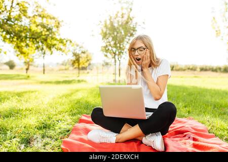 Emotional schönes Mädchen mit Brille, Blick auf den Laptop-Bildschirm mit großer Bewunderung, sitzt auf einer Decke im Park Stockfoto