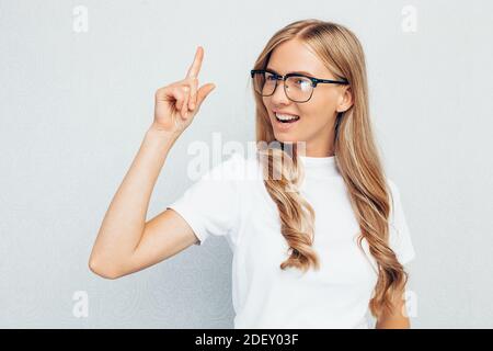 Schöne Studentin mit Brille, in weißem T-Shirt, sitzt auf dem Boden, eine Idee und Blick auf die Kamera auf einer grauen Wand Hintergrund Stockfoto