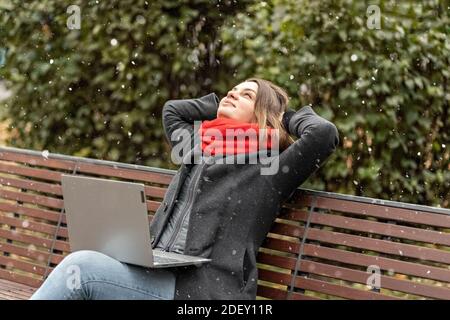 Eine junge Frau, mit den Händen hinter dem Kopf, fühlt Ruhe, ruht nach der Arbeit, sitzt auf einer Holzbank im Park ein Laptop auf den Knien. Pause während w Stockfoto