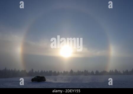 Oberwiesenthal, Deutschland. Dezember 2020. Ein Auto fährt über einen verschneiten Parkplatz, während im Hintergrund die Sonne durch den Eisnebel einen Heiligenschein erzeugt. Das Licht wird durch die Eiskristalle gebrochen und die typischen Ringe oder andere Formen entstehen. Quelle: Jan Woitas/dpa-Zentralbild/dpa/Alamy Live News Stockfoto