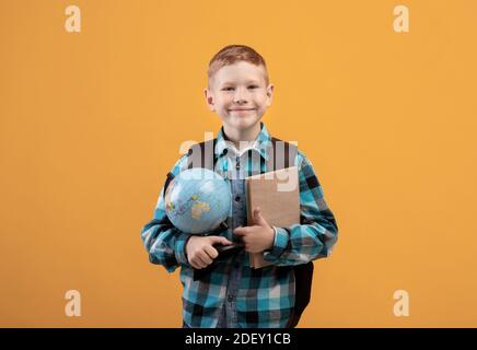 Positive Schuljunge mit Rucksack hält Haufen von Büchern und Globus Stockfoto