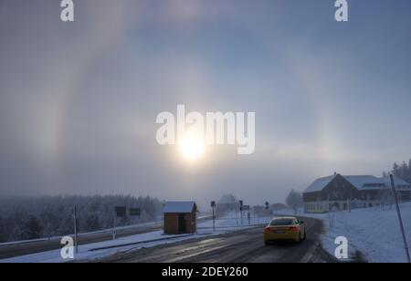 Oberwiesenthal, Deutschland. Dezember 2020. Ein Auto fährt über eine verschneite Straße, während im Hintergrund die Sonne durch den Eisnebel einen Heiligenschein erzeugt. Das Licht wird durch die Eiskristalle gebrochen und die typischen Ringe oder andere Formen entstehen. Quelle: Jan Woitas/dpa-Zentralbild/dpa/Alamy Live News Stockfoto