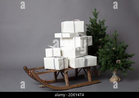 Rustikaler Schlitten mit ein paar weiß verpackten Geschenkboxen neben zwei Miniatur-Weihnachtsbäumen. Horizontales Bild auf grauem Hintergrund mit Kopierbereich. Stockfoto