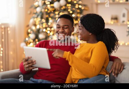Happy Millennial Dame zeigt Finger auf Tablette und zeigt Guy etwas Interessantes im Inneren mit Baum und Girlanden Stockfoto