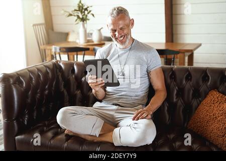 Glücklicher Mann mittleren Alters in legerer Kleidung mit Videoanruf Auf einem digitalen Tablet, während Sie in Lotuspose auf dem Sofa sitzen Im Wohnzimmer Stockfoto