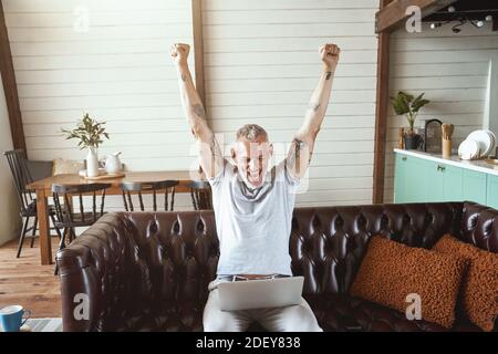 Erfolg wird gefeiert. Aufgeregter Mann mittleren Alters in legerer Kleidung, der auf den Laptop-Bildschirm schaut und die Hände hochhebt, während er auf dem Sofa im Wohnzimmer sitzt Stockfoto