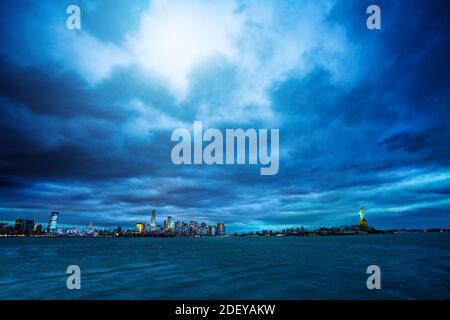 Breites dramatisches Panorama der Freiheitsstatue über dem New York Stadtbild in der Abenddämmerung und bewölktem Wetter Stockfoto