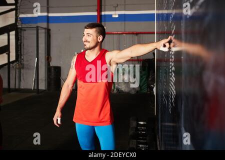 Stock Foto von einem kaukasischen Mann zeigt auf Trainingspläne geschrieben auf einer Kreidetafel. Er lächelt. Er trägt ein rotes Tanktop und eine blaue Hose. Stockfoto