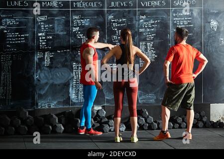 Stock Foto von hinten von Baum Menschen überprüfen Trainingspläne auf einem Schwarzen Brett. Es gibt zwei Männchen und ein Weibchen. Sie sind nicht erkennbar. Stockfoto