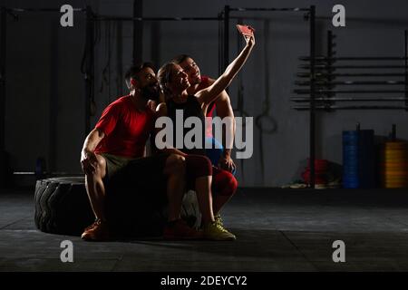 Stock Foto von drei Erwachsenen in einem Fitnessstudio sitzen auf einem Traktor Reifen. Sie machen ein Selfie und lächeln.Sie tragen Sportkleidung. Stockfoto