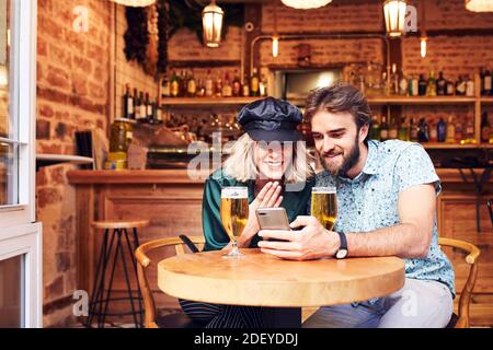 Stock Foto von einem Paar in ihren 30ern einen Drink an einer Bar. Sie schauen sich ein Video auf seinem Smartphone an. Sie tragen legeres Tuch. Stockfoto