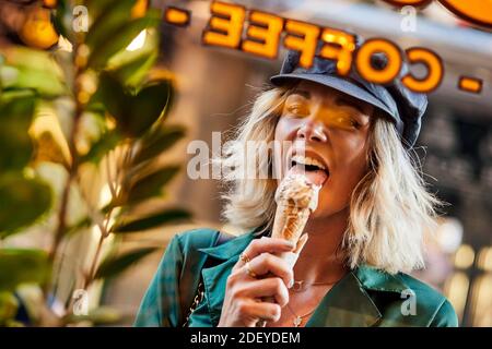 Stock Foto von einer blonden Frau in ihren 30ern durch ein Fenster beim Eislecken schauen. Sie trägt ein legeres Tuch und einen Hut. Stockfoto