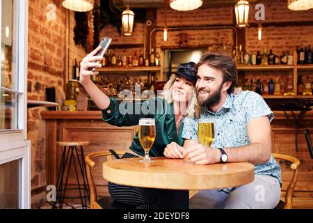 Stock Foto von einem Paar in ihren 30ern einen Drink an einer Bar. Sie machen ein Selfie. Sie tragen legeres Tuch. Stockfoto