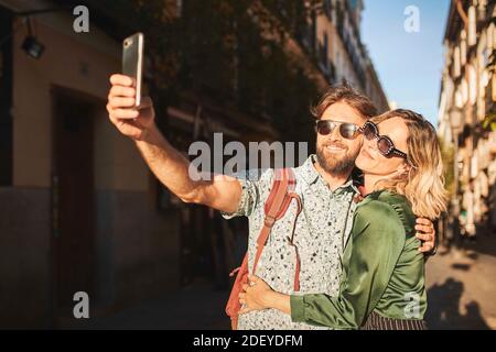 Stock Foto von einem Paar in ihren 30ern zu Fuß eine Straße hinunter. Sie machen ein Selfie. Sie tragen legeres Tuch. Stockfoto