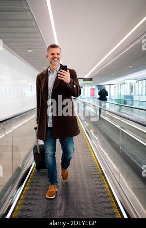 Stock Foto von einem kaukasischen Geschäftsmann, der auf einem Förderband mit einem Koffer geht, um einen Flug zu nehmen. Er lächelt und schaut auf sein Telefon. Er Stockfoto