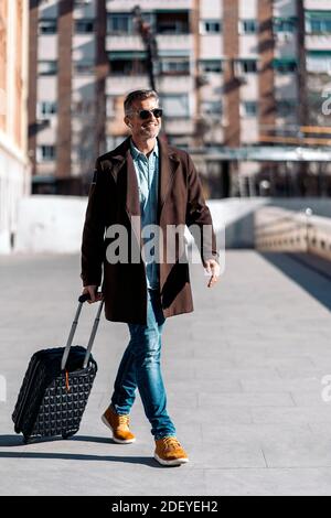 Stock Foto von einem kaukasischen Business Casual Mann zu Fuß in der Straße. Er lächelt. Er trägt Freizeitkleidung und Sonnenbrille. Er hat einen Koffer. Stockfoto