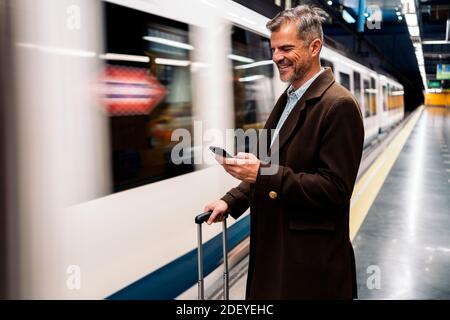 Stock Foto von einer Seitenansicht eines Geschäftsmanns tippen in seinem Smartphone wartet auf die U-Bahn in der Plattform nehmen. Er lächelt und trägt Casua Stockfoto